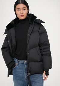 Jachetă puffer neagră cu guler din blană artificială, închidere cu fermoar și capse, având secțiuni mari matlasate și tiv ajustabil, asociată cu un pulover negru cu guler înalt.
