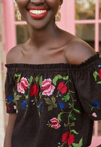 Black, off-the-shoulder top with colorful floral embroidery in red, pink, blue, and green. Ruffled neckline and textured fabric.