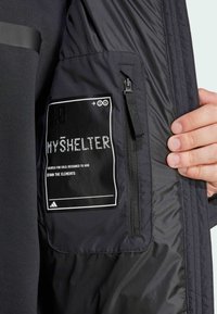 Crna jakna glatke teksture sadrži džep s vidljivom etiketom na kojoj piše "MYSHELTER" i zatvaračem, dizajnirana za hladno vreme.