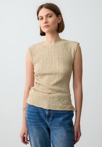 Beige getextureerde mouwloze top met een slim fit en subtiele plooien, gecombineerd met lichtblauwe rechtlopende jeans.