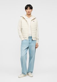 Άνδρας που φοράει εκρού κοντή γιλέκο με επένδυση πάνω από μπλούζα με μακριά μανίκια, ανοιχτό μπλε τζιν παντελόνι φαρδιάς γραμμής και λευκά αθλητικά παπούτσια, στέκεται μπροστά σε απλό φόντο.