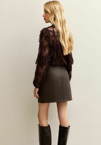Mini gonna in pelle marrone con texture abbinata a una blusa trasparente scura con dettagli arricciati, vista da dietro. I capelli lisci scendono.