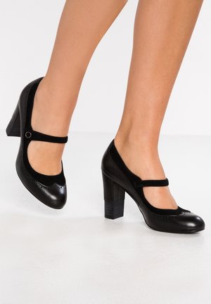Femme portant des chaussures Mary Jane en cuir noir à talons blocs, un pied légèrement levé au-dessus d'un sol blanc.