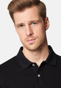 Czarna koszulka polo z kołnierzykiem i dwoma guzikami, wykonana z fakturowanej tkaniny. Zbliżenie na twarz mężczyzny z jasnoniebieskimi oczami i krótkimi włosami.