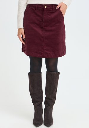 Femme portant une jupe en velours côtelé bordeaux avec des poches, des collants noirs et des bottes en daim brun foncé montant jusqu’au genou sur un fond uni.