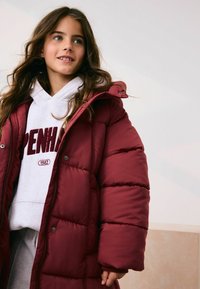 Burgunderfarvet puffjakke med hætte, der har kviltet tekstur og trykknapper, lagdelt over en grå sweatshirt med teksten 'Copenhagen'.