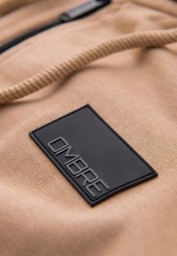 Patch rectangulaire noir avec texte en relief "OMBREE" sur tissu beige avec couture froncée et cordon beige.