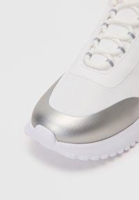 Λευκό αθλητικό παπούτσι με λείο ασημί καπάκι στην μύτη, υφή στο πάνω μέρος με ανακλαστικές λεπτομέρειες και λευκή καουτσούκ σόλα.