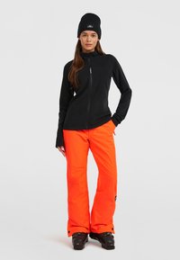 Zwarte fleece jas met een rits, gecombineerd met feloranje skibroek en zwarte sneeuwlaarzen. Beanie inbegrepen, alles in een minimalistische stijl.