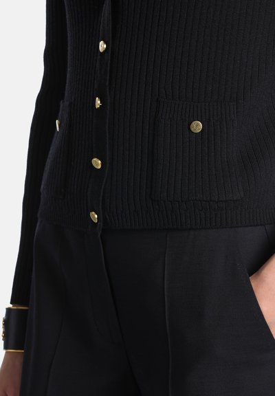 Cardigan noir côtelé avec boutons dorés et détail de poche, porté avec un pantalon noir et un bracelet noir et doré au poignet.