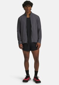Young man standing wearing a grey zip-up jacket, black shorts, black socks, and red and black sneakers on a white background.