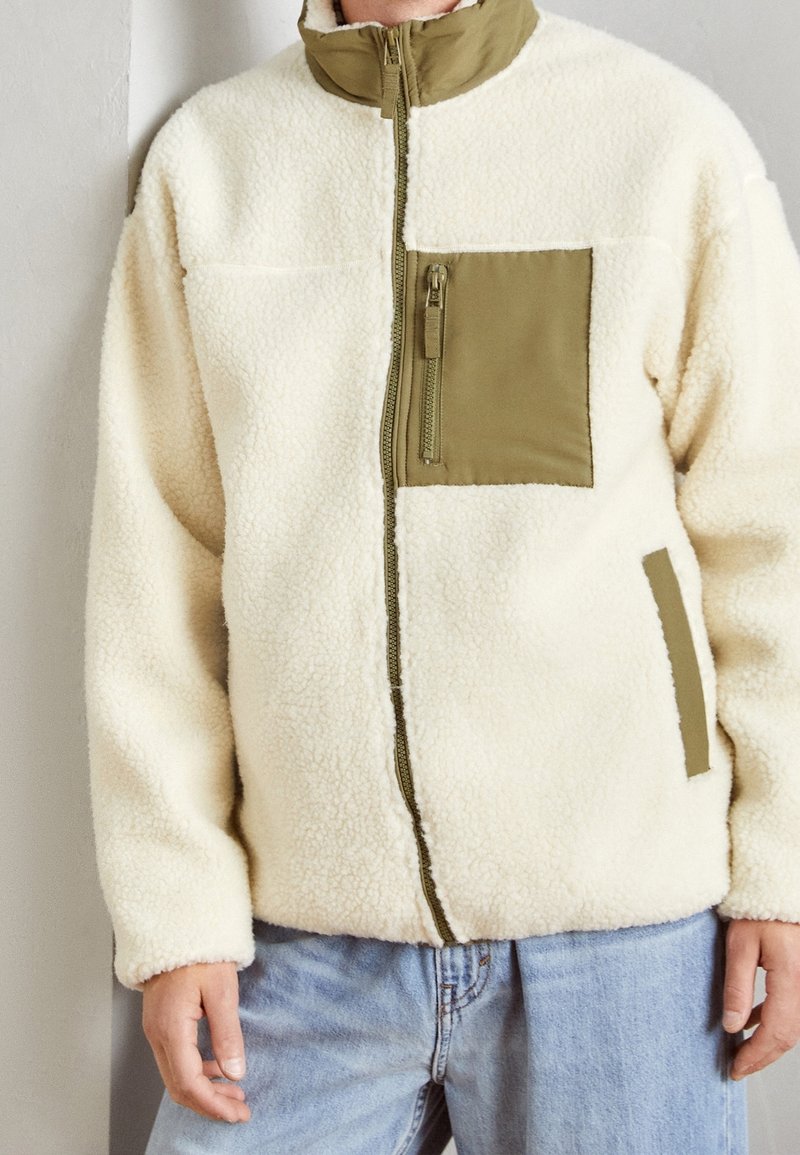 Persoon die een crèmekleurig fleecejack draagt met een olijfgroene, met ritssluiting afgesloten borstzak en lichtblauwe spijkerbroek, staand binnen.