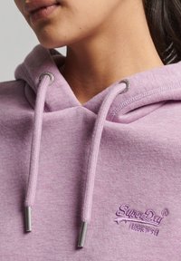 Sudadera de color púrpura claro con una textura suave, que cuenta con cordones con puntas plateadas y el logo bordado "Superdry" en un color a juego en la parte delantera.