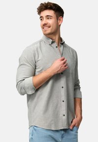 Camisa abotonada gris claro con un bolsillo, mangas largas arremangadas hasta los codos, botones verdes y una textura de tela suave.