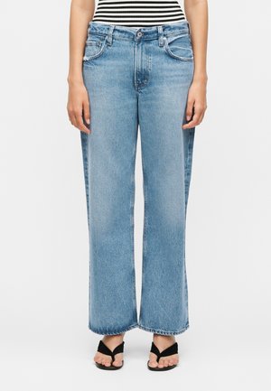 Személy világoskék egyenes szárú farmert, fekete flip-flop papucsot és fekete-fehér csíkos felsőt visel, sima háttér előtt áll.