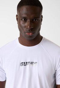 Weißes, kurzärmeliges Sportshirt mit Mesh-Textur und metallischem "GUESS"-Logo auf der Brust. Einfaches und modernes Design.