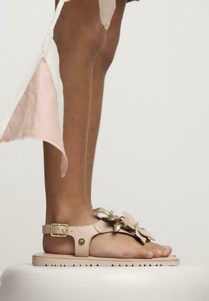 Sandalia beige con suela plana, acentos florales y tira en el tobillo. Fabricada con material sintético con superficie texturizada y hebilla ajustable.