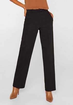 Femme portant un pantalon noir taille haute à jambes droites avec poches latérales, associé à des bottines marron à talons et un top marron rentré à l’intérieur.
