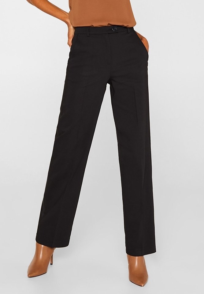 Femme portant un pantalon noir taille haute à jambes droites avec poches latérales, associé à des bottines marron à talons et un top marron rentré à l’intérieur.