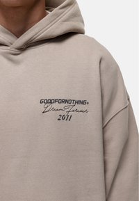 Felpa beige in cotone misto con tasca frontale a kanga. Sulla parte sinistra del petto c'è del testo ricamato in nero: "GOODFORNOTHING® Dream Followed 2011."