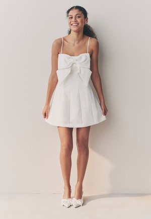 Ung kvinde smilende, iført hvid ærmeløs kjole med stor sløjfe foran og matchende hvide spidse hæle, står mod en neutral væg.