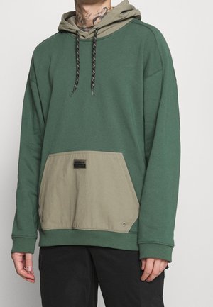 Homme portant un sweat à capuche vert avec une poche avant beige et une capuche, des cordons noirs à motifs, et un pantalon noir, debout devant un fond blanc.