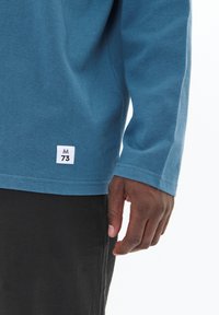 Blaues Langarmshirt aus Baumwolle mit figurbetontem Schnitt, Rundhalsausschnitt und einem kleinen weißen Etikett am Saum, auf dem "M 73" gedruckt ist.