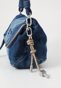 Denimová taška přes rameno v tmavě modré barvě se stříbrným zipem a kováním. Obsahuje uzlový béžový akcent na boku a má zaoblený tvar.