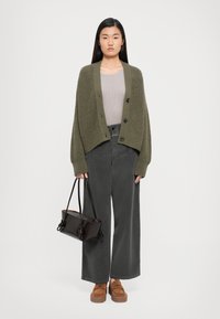 Mujer de pie con un cárdigan verde oliva de gran tamaño, blusa beige, pantalones anchos gris oscuro, mocasines marrones y sosteniendo un bolso marrón oscuro.