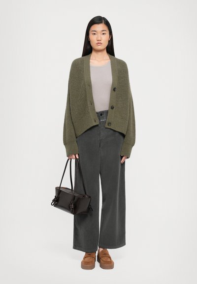 Femeie în picioare, purtând un cardigan supradimensionat de culoare verde măsliniu, un top bej, pantaloni largi de culoare gri închis, loafers maro și ținând o geantă maro închis.