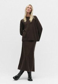 Bruine gebreide trui met een oversized pasvorm en geribbelde zoom, gecombineerd met een bijpassende maxi-jurk, gedragen met zwarte enkellaarsjes. Soepele textuur.