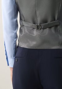 Back view of a person wearing a gray waistcoat with adjustable buckle and navy blue trousers paired with a light blue long sleeve shirt.