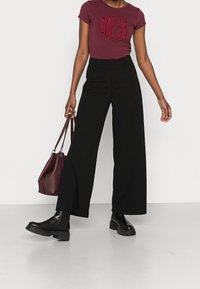 Kvinde iført en burgunder GUESS t-shirt, sorte brede bukser, sorte ankelstøvler, som holder en burgunder læderhandbag, stående på grå gulv.