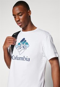 Tricou alb din bumbac, cu un logo Columbia colorat și diverse modele. Mâneci scurte și guler rotund. Proiectat pentru purtare casuală.