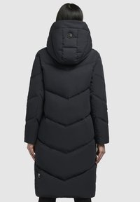 Sort polstret jakke med en chevron-mønster, en stor hætte og en cirkulær logo-detalje på ryggen. Designet er langt og tætsiddende.