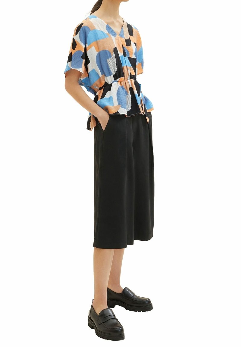 Top colorato e morbido con motivi astratti blu, arancioni e neri. Abbinato a culotte nere a gamba larga e mocassini neri.