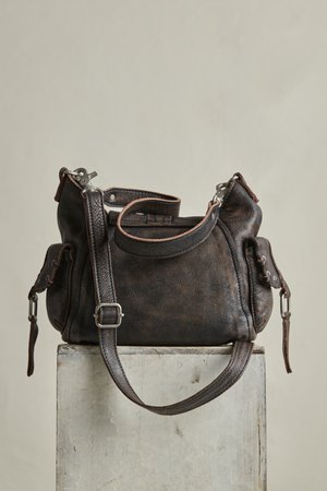 Temno rjava usnjena torbica z nastavljivim naramnim trakom in stranskimi žepi, nameščena na svetlo obarvan lesen blok na enostavni podlagi.