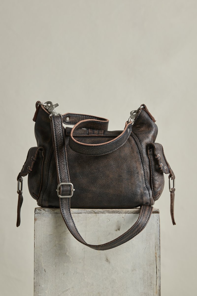 Σκούρα καφέ δερμάτινη τσάντα χειρός με ρυθμιζόμενο ιμάντα ώμου και πλαϊνές τσέπες, τοποθετημένη σε ανοιχτόχρωμο ξύλινο τεμάχιο πάνω σε απλό φόντο.