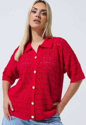 Kvinde iført en knaldrød, kortærmet, knappet strikbluse med geometriske hulmønstre, parret med lyseblå denimjeans.