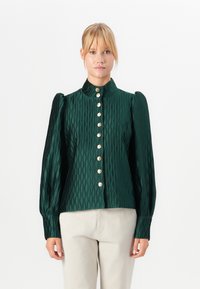 Woman wearing a dark green textured blouse with puffed long sleeves and gold buttons, paired with light beige pants, standing against white background.