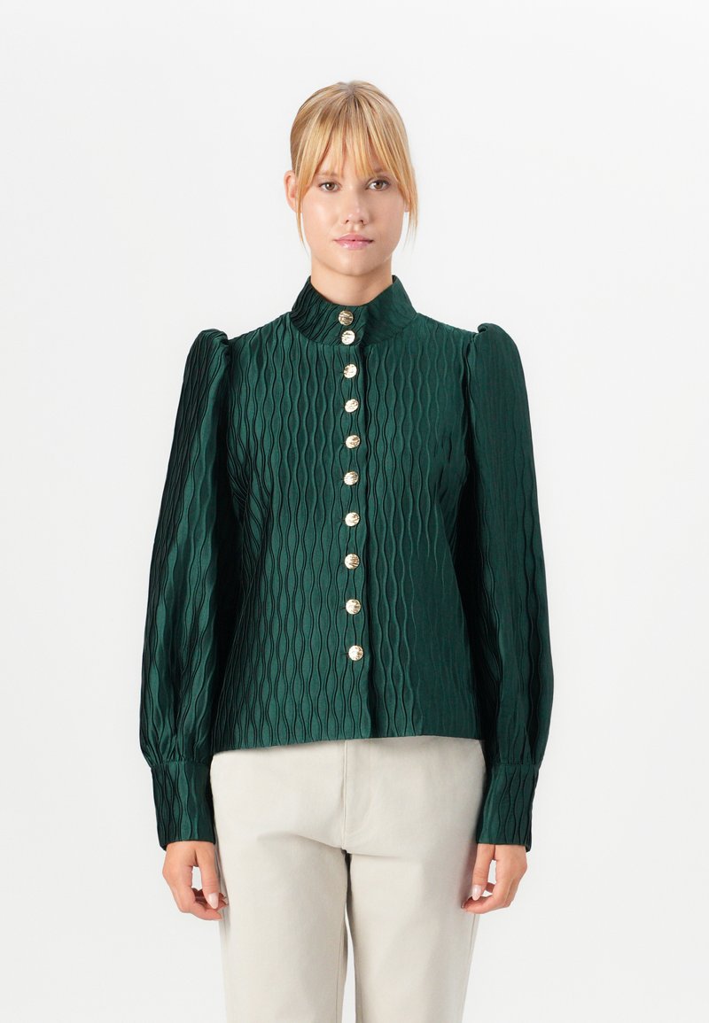 Mulher vestindo uma blusa texturizada verde escura com mangas longas bufantes e botões dourados, combinada com calças bege claras, encostada a um fundo branco.