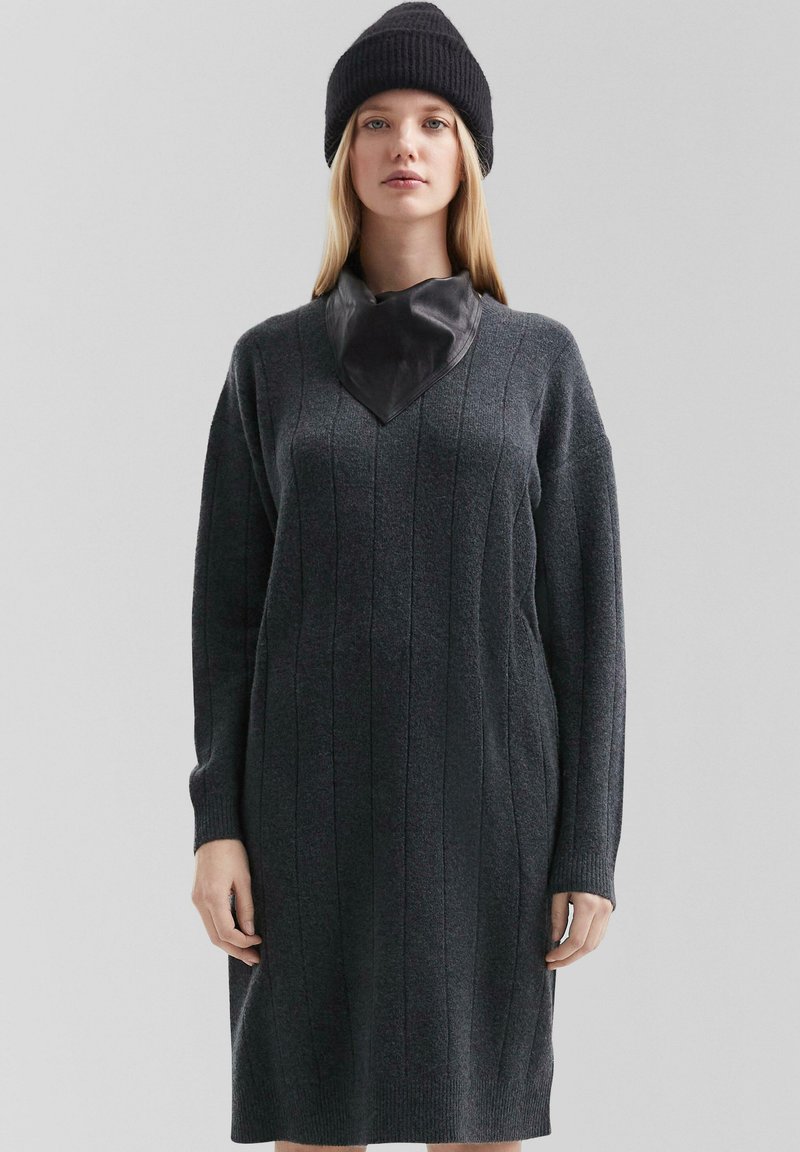 Anthrazitgraues Strickkleid mit langen Ärmeln, rippstrukturiert und mit einem V-förmigen Lederkragen. Kombiniert mit einer schwarzen Strickmütze.