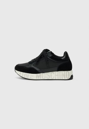 Zwarte sneakers gemaakt van een combinatie van leer en suede, met een dikke witte zool waarop "Moschino" in grijze letters is weergegeven.