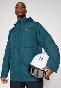 Veste imperméable bleu-vert avec de multiples poches, fermeture éclair et capuche ajustable. Casque blanc tenu à la main, comportant deux fentes de ventilation.