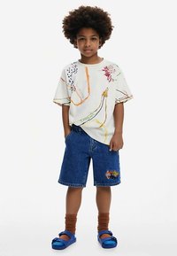 Bijela prevelika majica s šarenom apstraktnom vezom, uparena s plavim traper šortsevima s šarenim zakrpama, smeđim čarapama i plavim sandalama.