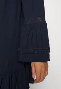 Vestido navy con mangas largas de campana, adornado con encaje en el dobladillo y un volante. Tejido suave y detalles texturizados adornan el diseño.