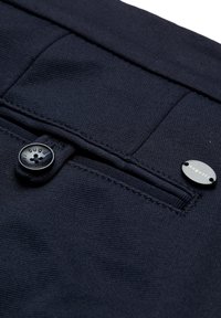 Tissu bleu marine foncé avec une poche à rabat boutonnée et une petite plaque métallique ovale portant la marque « bugatti » près du bord de la poche.