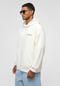 Crèmewitte hoodie met een gestructureerde stof, een voorzak en een capuchon met trekkoord. Bevat geborduurde tekst "Eternity Project" op de borst.