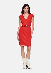 Rotes figurbetontes Kleid mit Kappenärmeln und V-Ausschnitt, akzentuiert durch einen kontrastierenden Gürtel und weißen Saumabschluss. Kombiniert mit weißen Knöchelstiefeln.