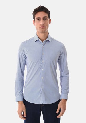 Uomo di fronte che indossa una camicia aderente a maniche lunghe con bottoni, a righe verticali bianche e azzurre, e pantaloni scuri.